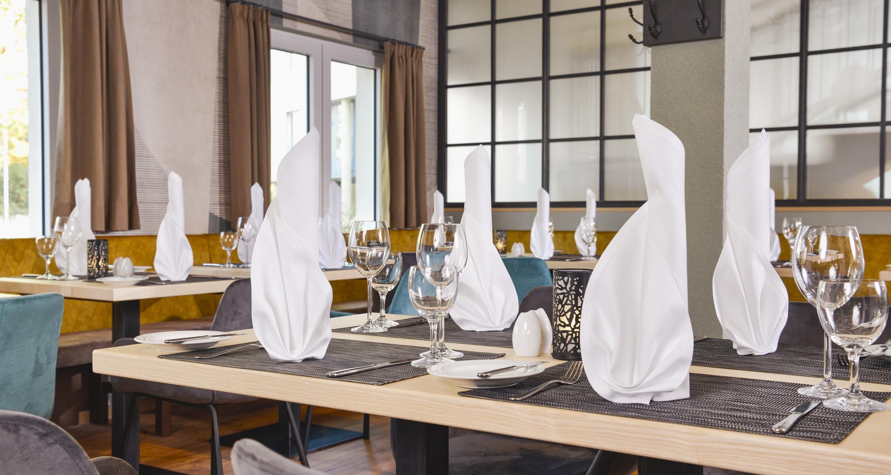 L’intérieur d’un restaurant, avec des tables dressées pour le service, où l’on peut admirer de la belle verrerie transparente et des serviettes blanches.