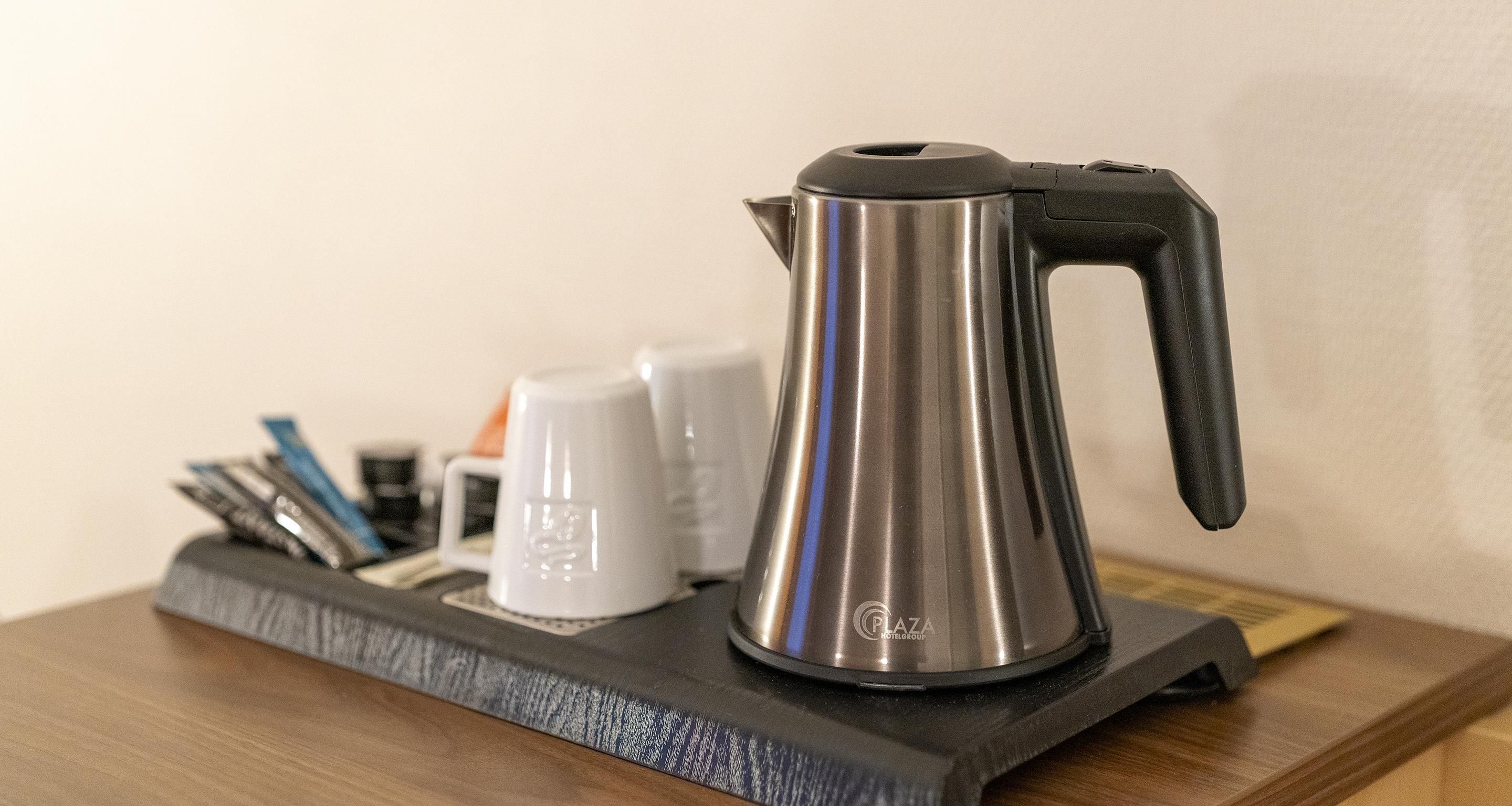 A metallic kettle on a wooden tray with a black handle, accompanied by white cups and a spoon set.