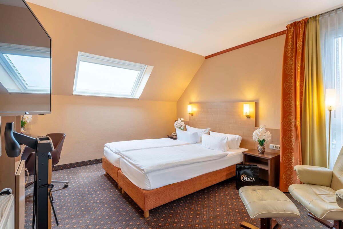 Une chambre d’hôtel avec un lit, un bureau, une chaise, une petite table ornée d’un vase de fleurs et une fenêtre avec des rideaux.
