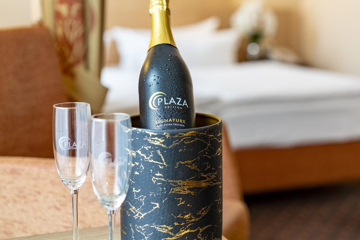 A bottle of champagne in a decorative holder with two glasses beside it, set on a table in a room with a bed and a nightstand in the background.