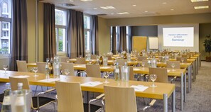 Ein Konferenzraum mit Seminartischen, ausgestattet mit Wasserflaschen, Weingläsern und einer Projektionsleinwand, die eine Willkommensbotschaft zeigt.