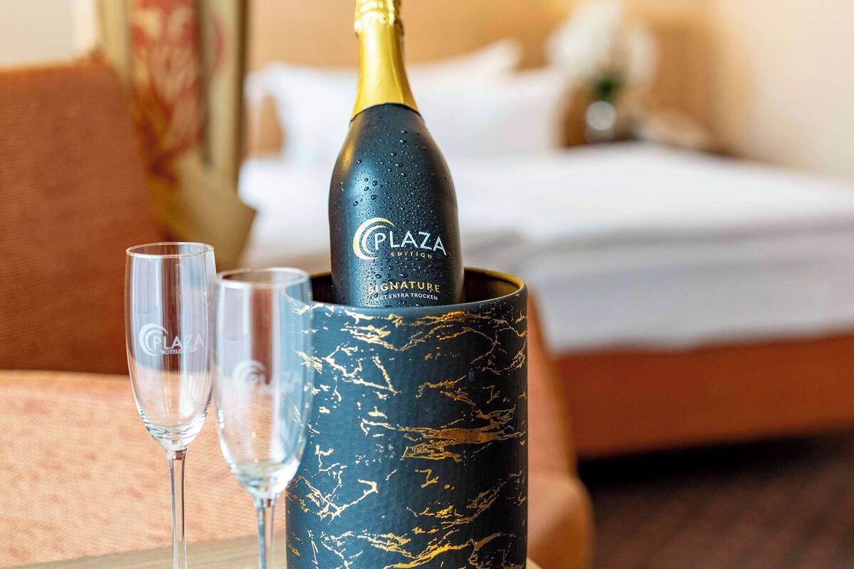 Een fles Plaza Signature Extra Trocken champagne in een decoratieve houder, met twee lege champagneglazen ernaast, op een tafel in een hotelkamer.