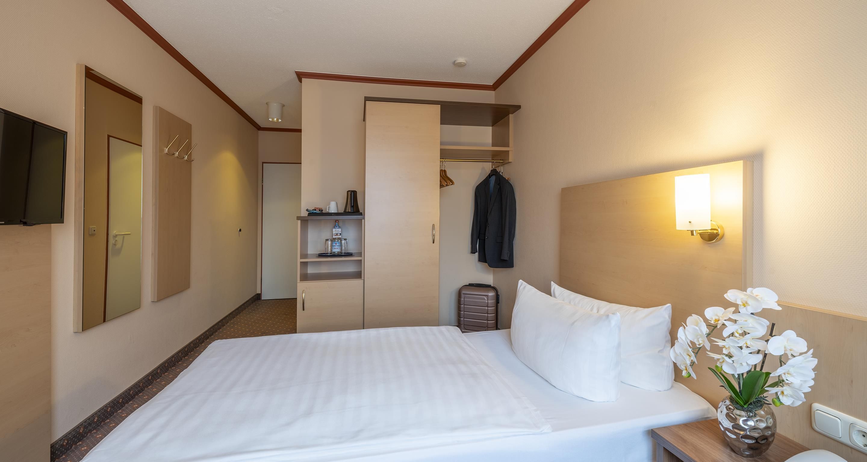A hotel room with a bed, a wardrobe, a mirror, a television, a bedside table with a vase of flowers, and a wall-mounted light.