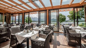 A restaurant with outdoor seating, wooden beams, and large windows overlooking a lake and mountains. - Hotel Am Hopfensee (Fuessen)