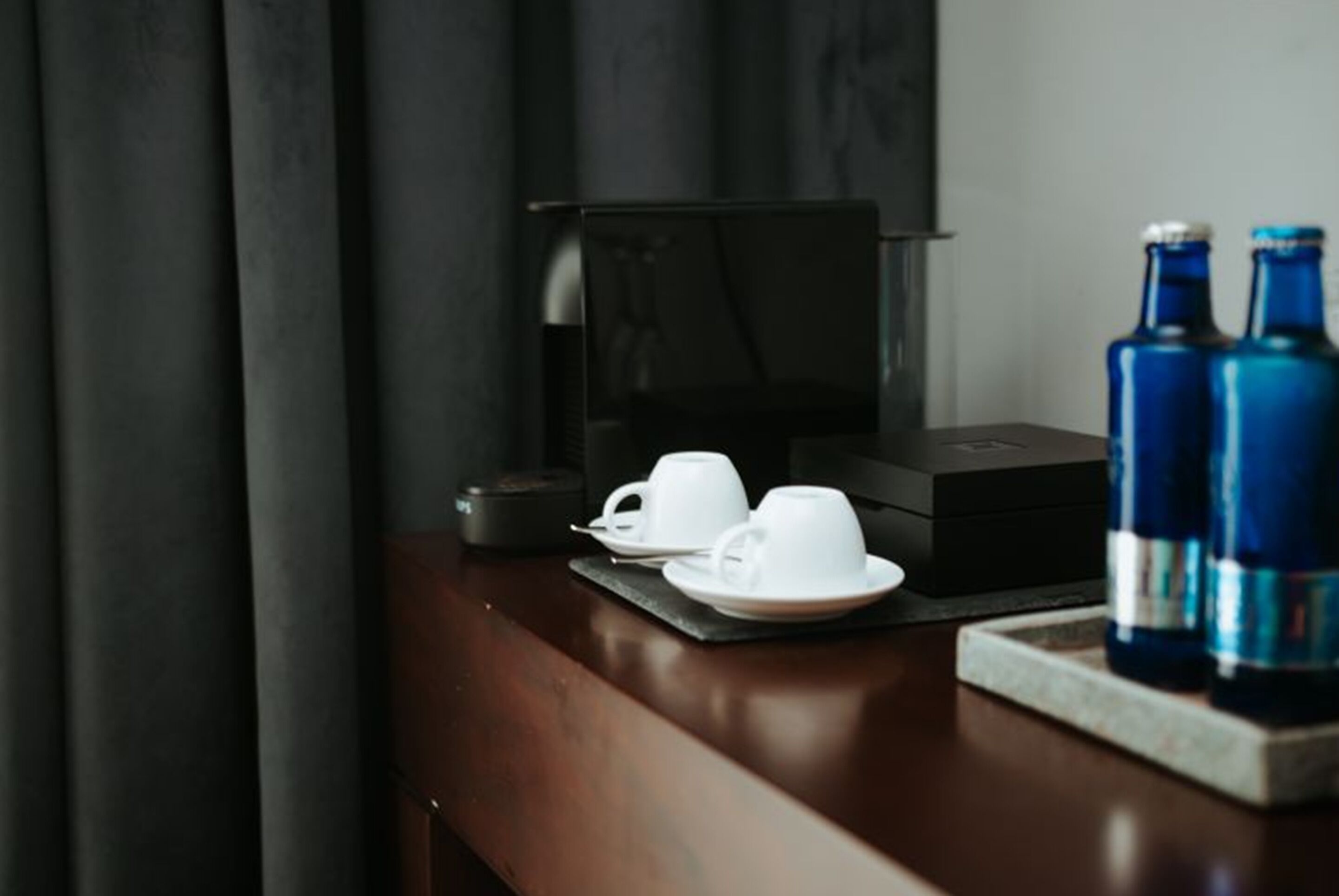 Una mesa de madera oscura con una cafetera, dos tazas blancas y dos botellas azules.