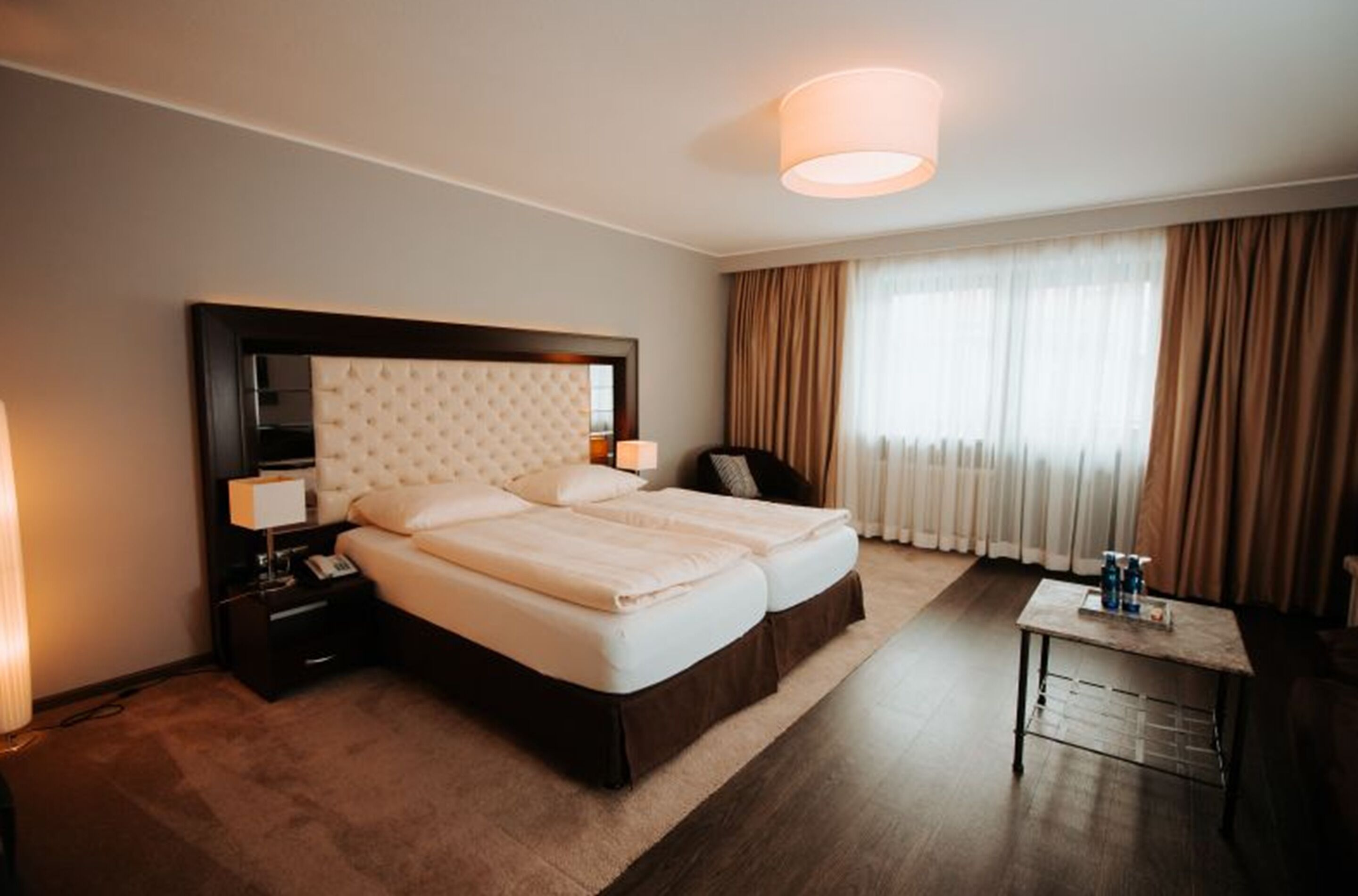 Habitación de hotel con una cama grande, una mesita de noche, una lámpara, una pequeña mesa con botellas y una ventana con cortinas.