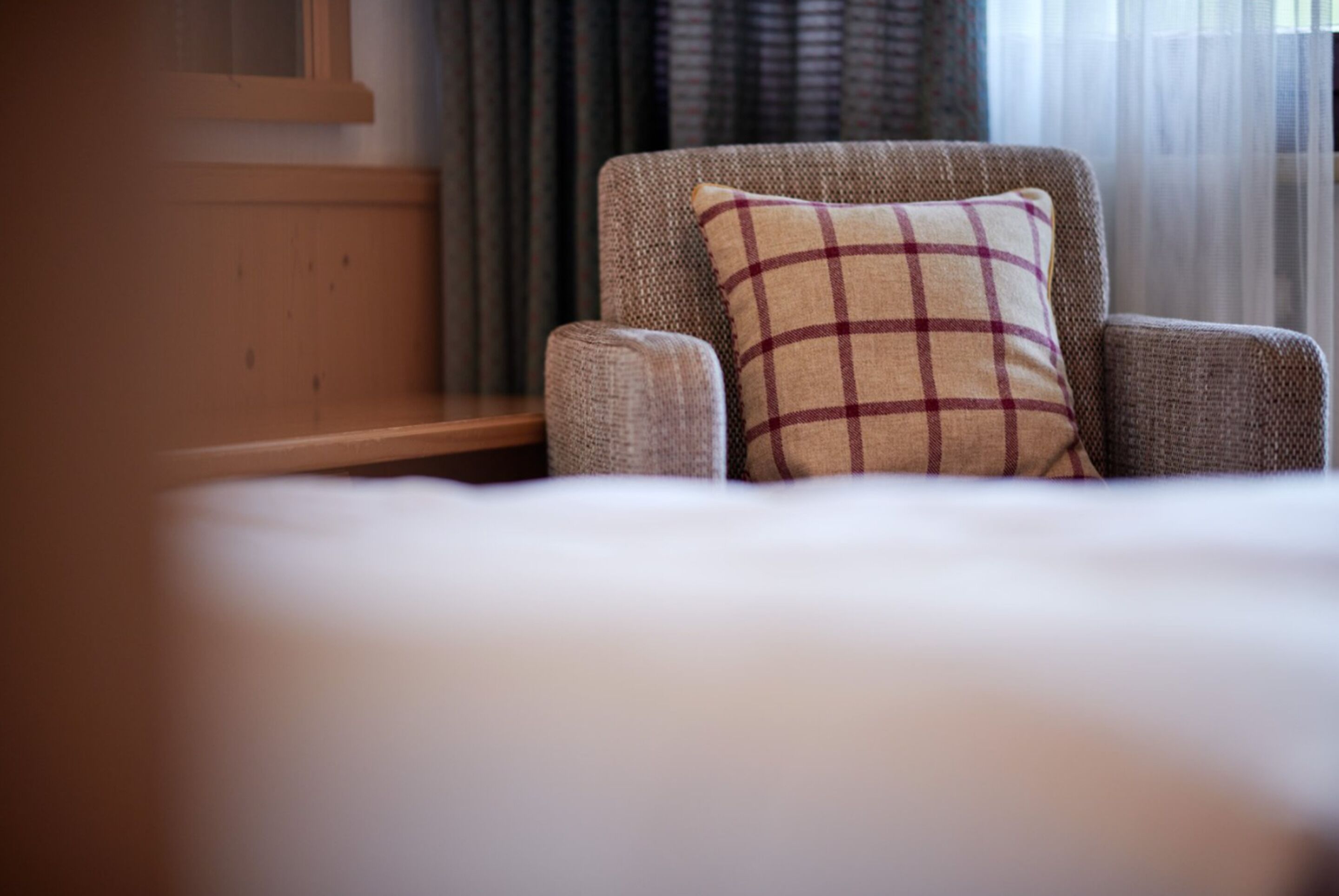 A hotel room with a bed, a chair with a plaid pillow, a wooden bench, and a window with curtains.