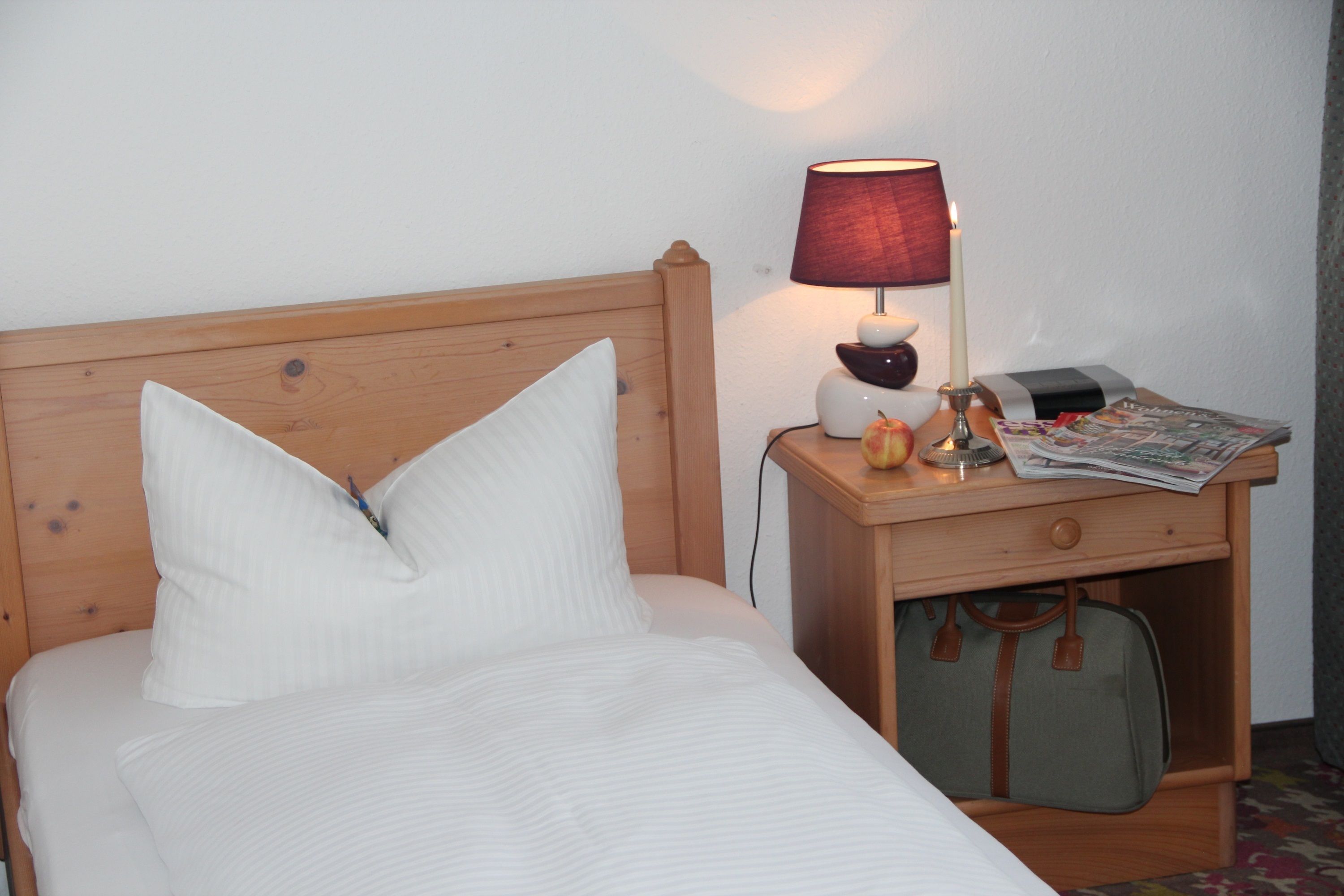Un lit bien fait, avec du linge de lit blanc, une tête de lit en bois, une table de chevet avec une lampe, une petite horloge, un magazine et une valise placée sous la table.