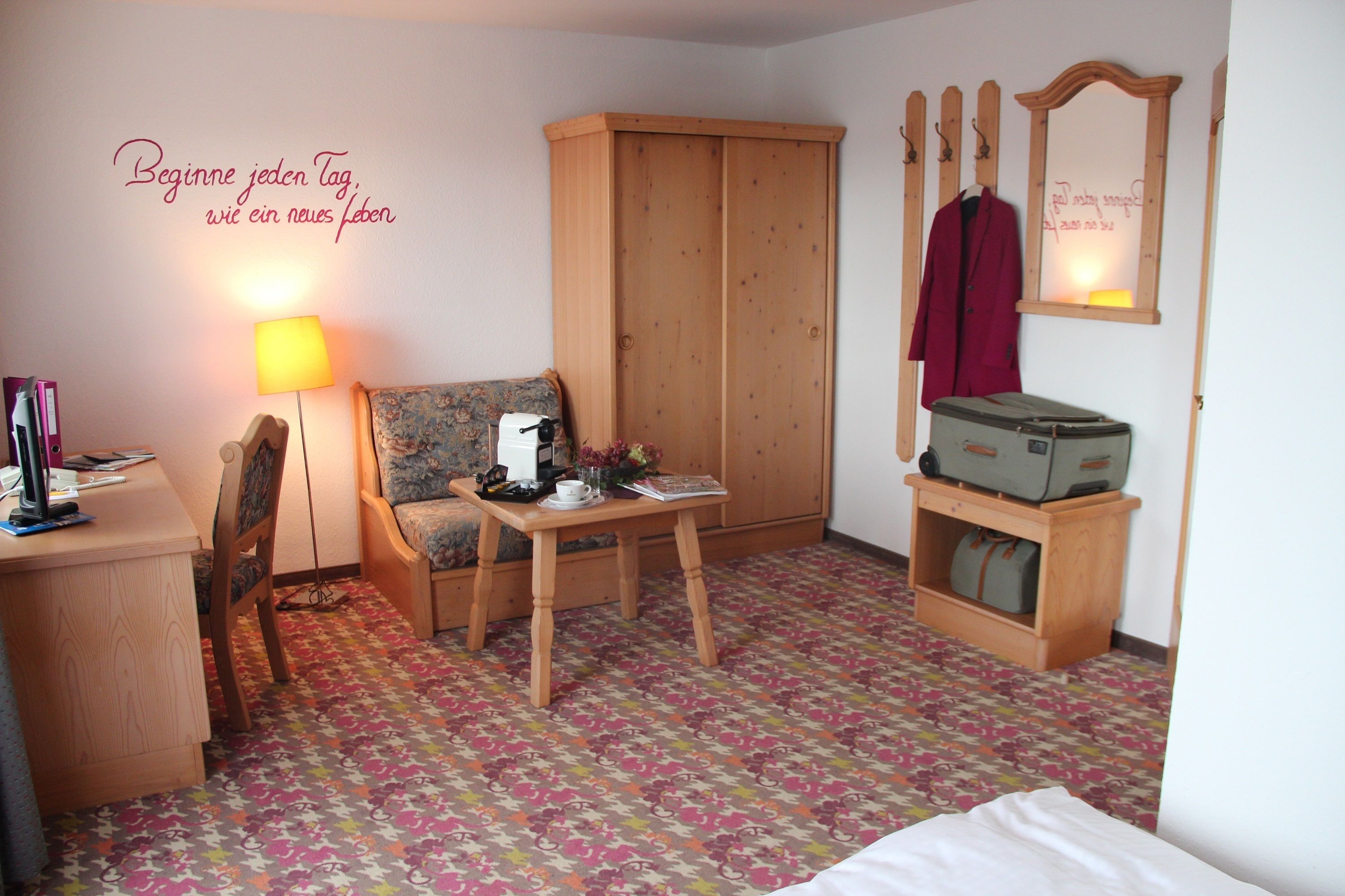 A cozy room with a wooden wardrobe, a sofa, a small table with a lamp, and a suitcase on a wooden cabinet.
