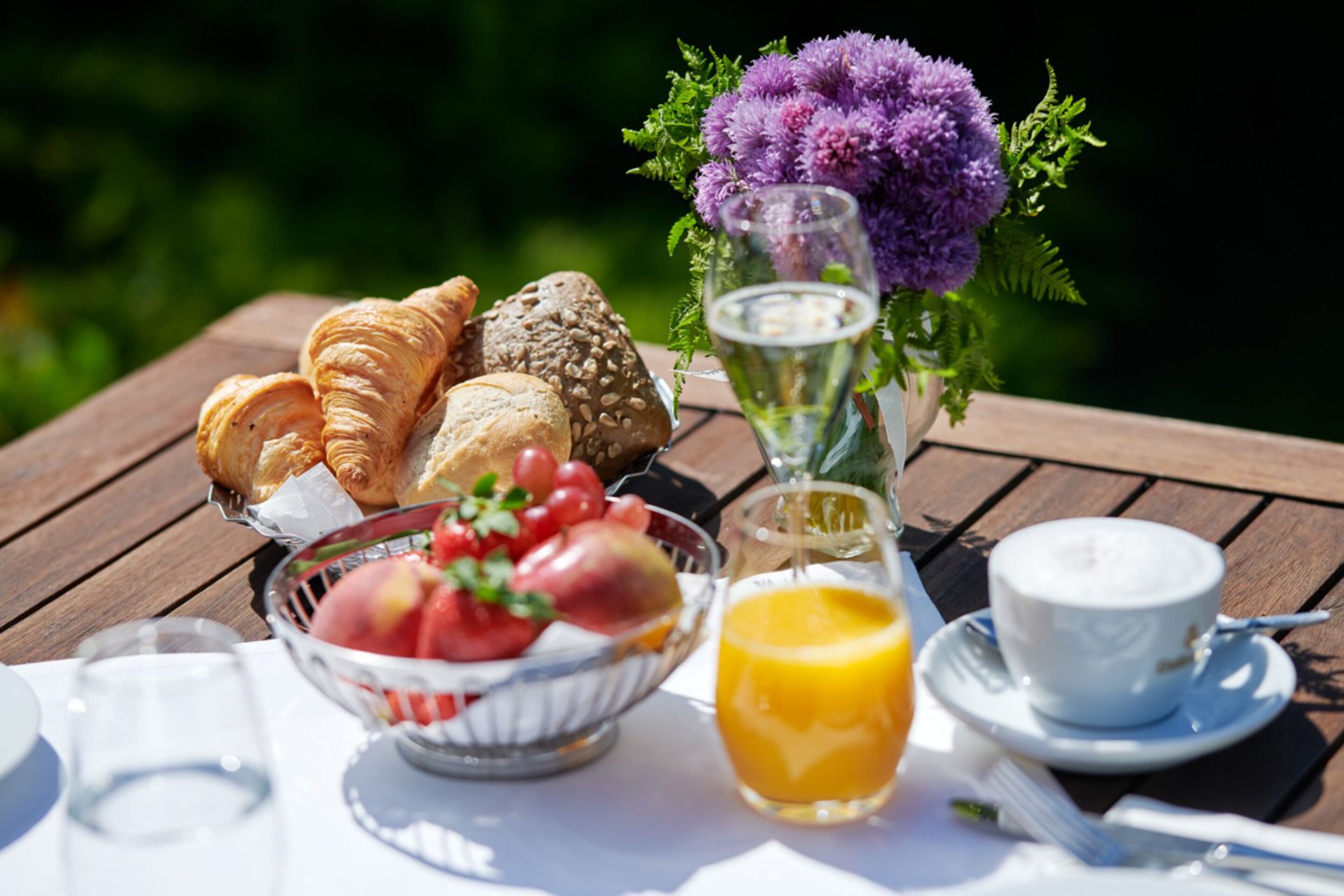 A table with croissants, a bowl of fruit, a glass of orange juice, a cup of coffee, and a vase with purple flowers.