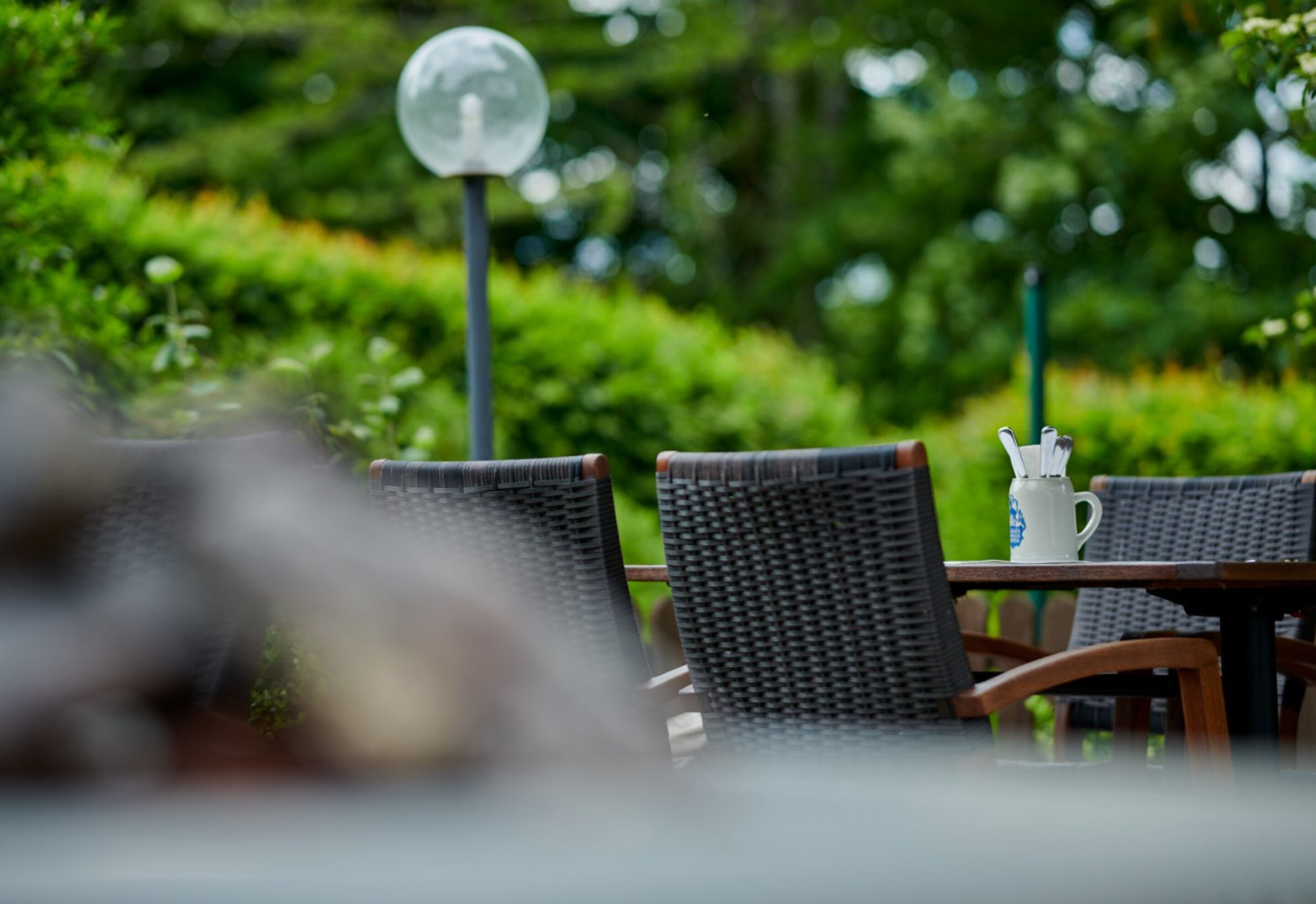 Een tafel met rieten stoelen en een witte kruik, in een tuin met een groene haag en een bolvormige lamp op de achtergrond.