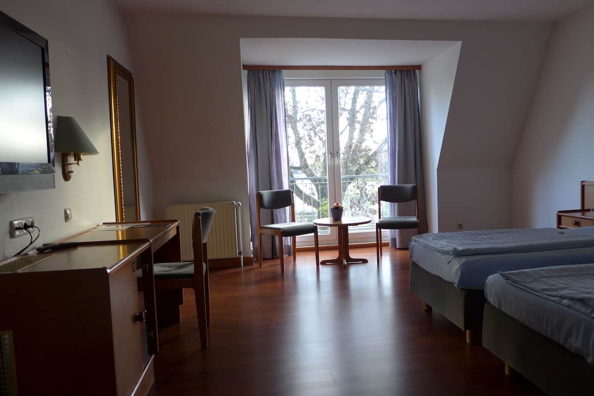 Quarto de hotel com duas camas, uma escrivaninha, uma cadeira, uma mesinha com uma planta e uma janela com cortinas.