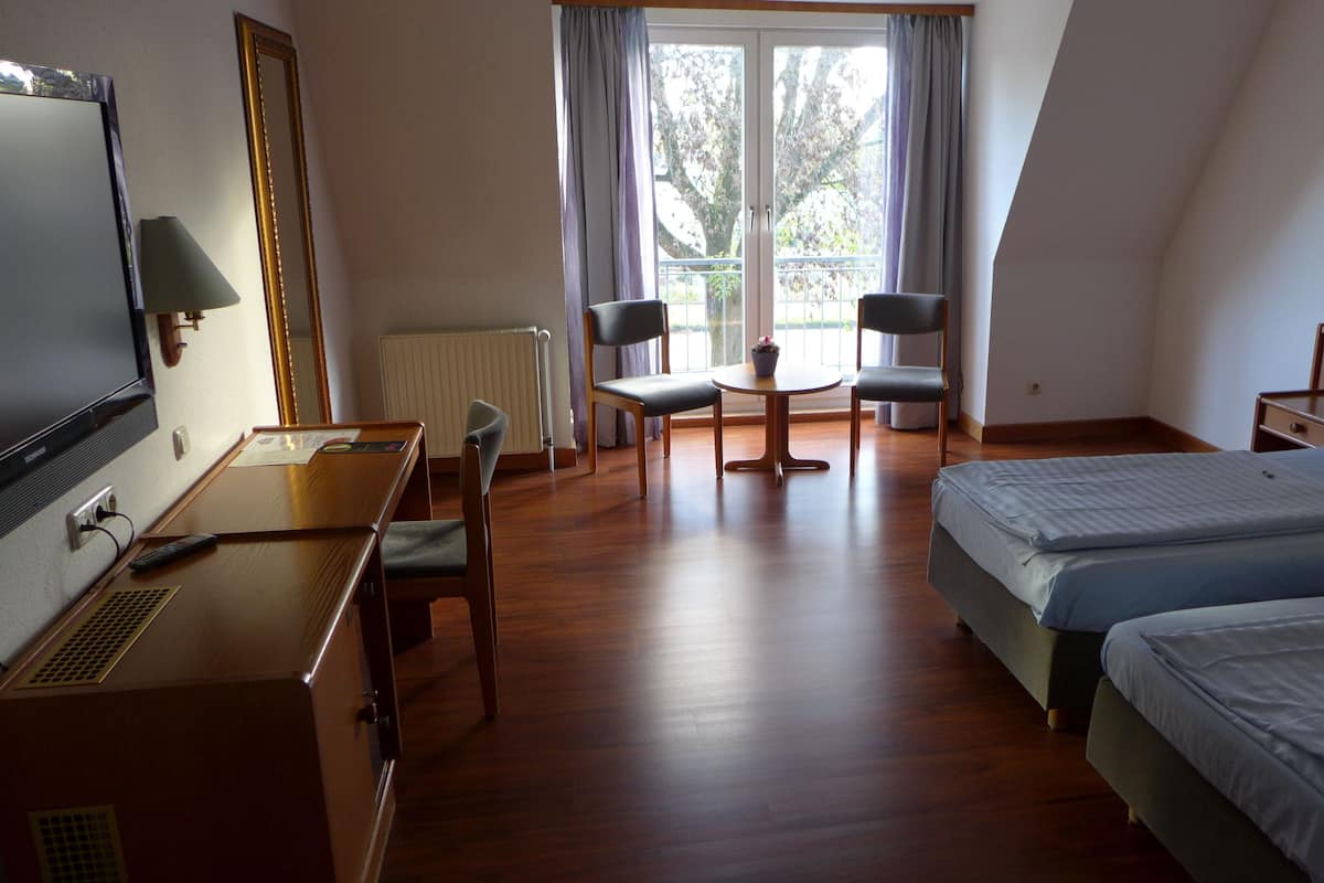 Quarto de hotel com escrivaninha de madeira, duas camas de solteiro, uma mesa pequena com cadeiras e uma janela grande com cortinas.