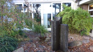 A garden with a variety of plants, a stone structure, and a building with large windows in the background. - Hotel Heidpark (Lueneburg)