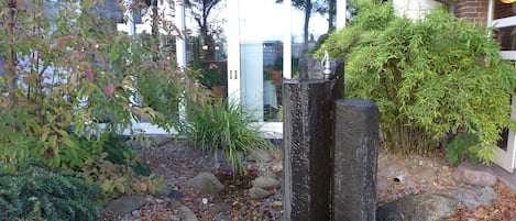 A garden with a variety of plants, a stone structure, and a building with large windows in the background.