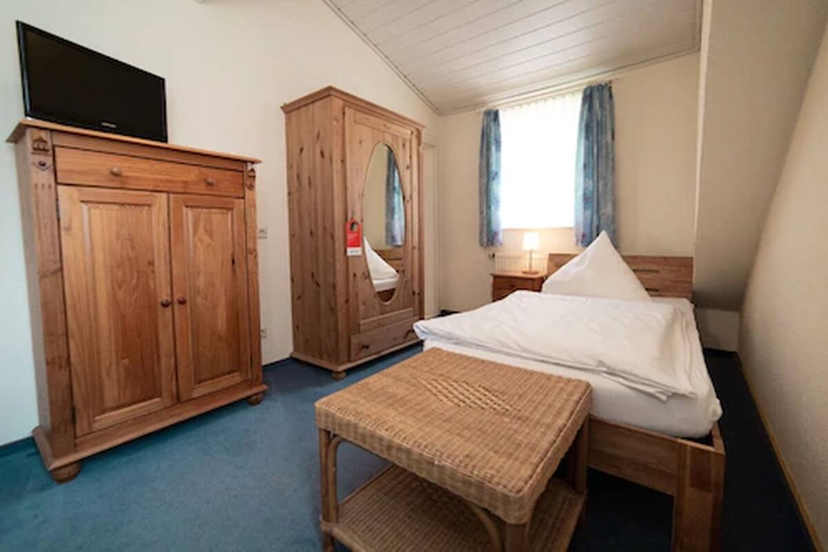 Une chambre à coucher avec un lit, une armoire en bois, un téléviseur posé sur l’armoire et une fenêtre avec des rideaux.