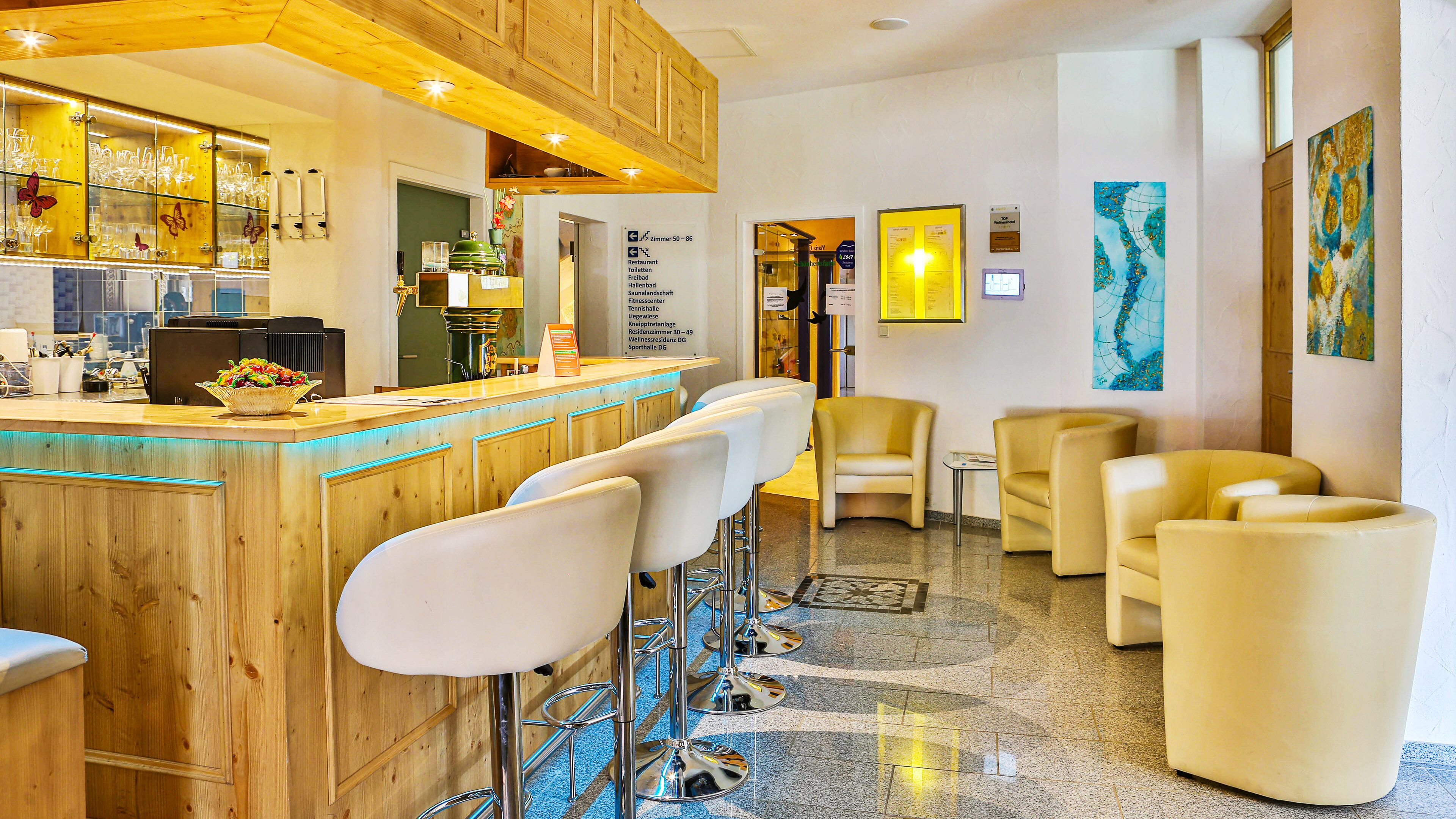 A bar area with wooden bar counter and white bar stools, a reception desk, and a seating area with yellow chairs.