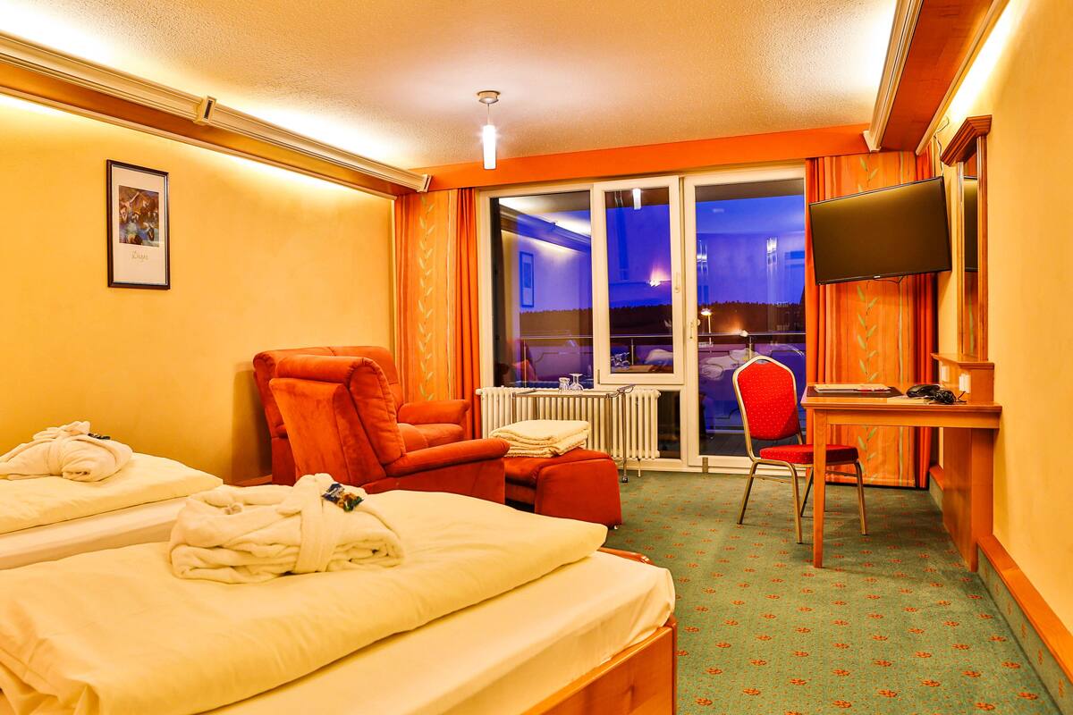 Une chambre d’hôtel avec deux lits, un fauteuil rouge, un bureau avec une chaise, une télévision et une grande fenêtre donnant sur l’extérieur.