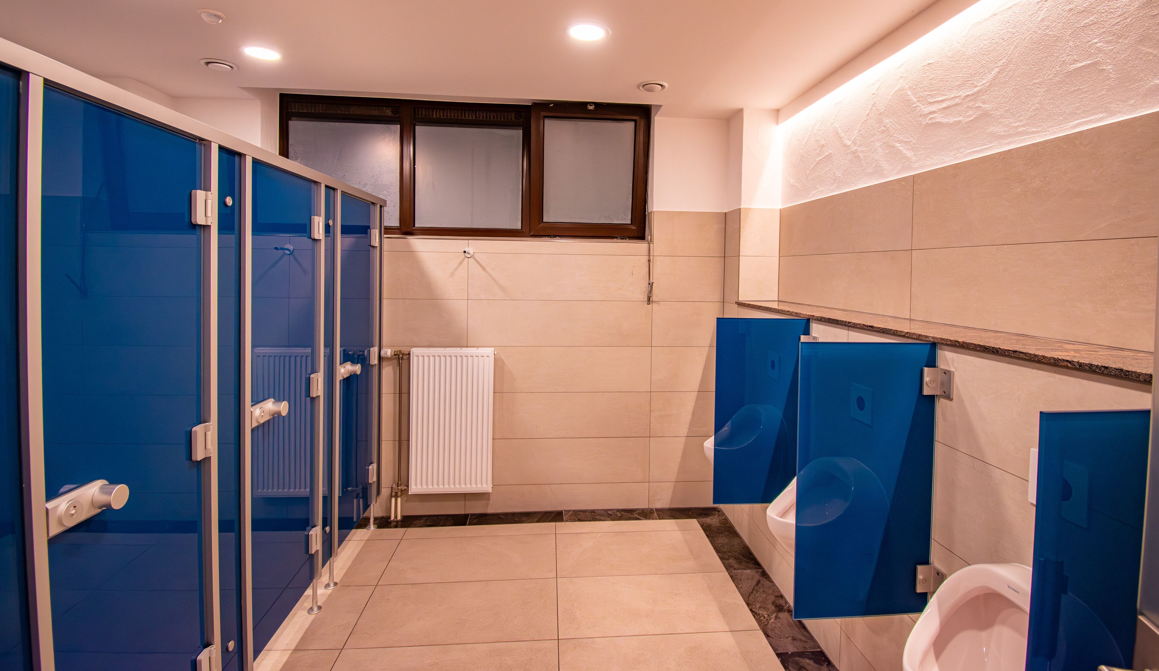 Un baño público con cabinas azules, orinales blancos y suelo de baldosas.