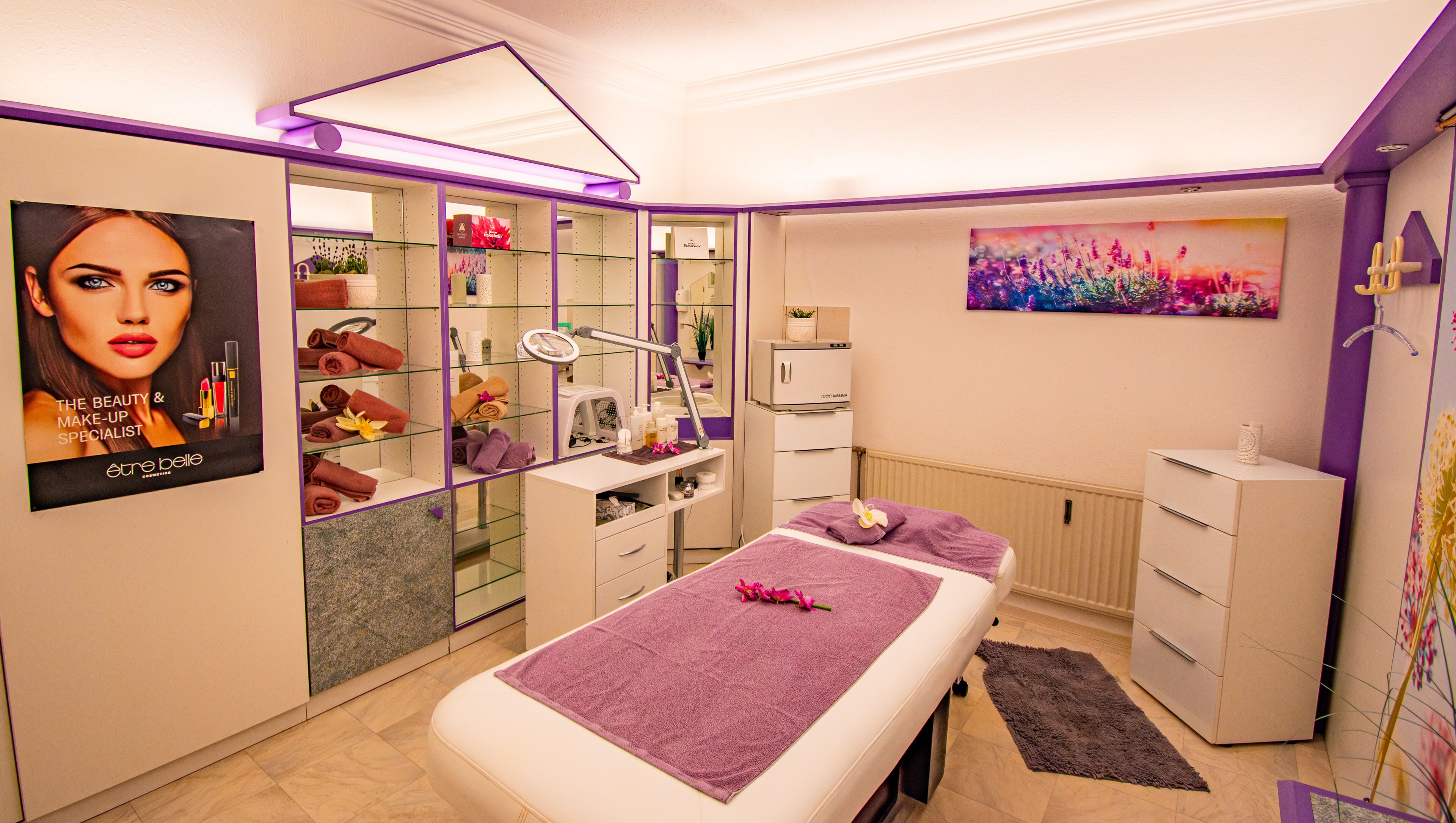 Een moderne spa ruimte met een massagetafel, planken met handdoeken, een wastafel met spiegel en een schilderij aan de muur.