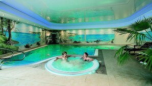 A pool area with a tropical mural, two people relaxing in the water, and a small hot tub. - Kur- und Wellness Hotel Mönchgut (Göhren)
