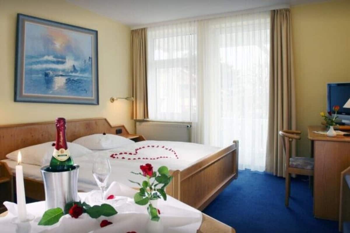 A hotel room with a bed, a bedside table with a bottle and glasses, a desk with a lamp, a painting on the wall, and a window with curtains.