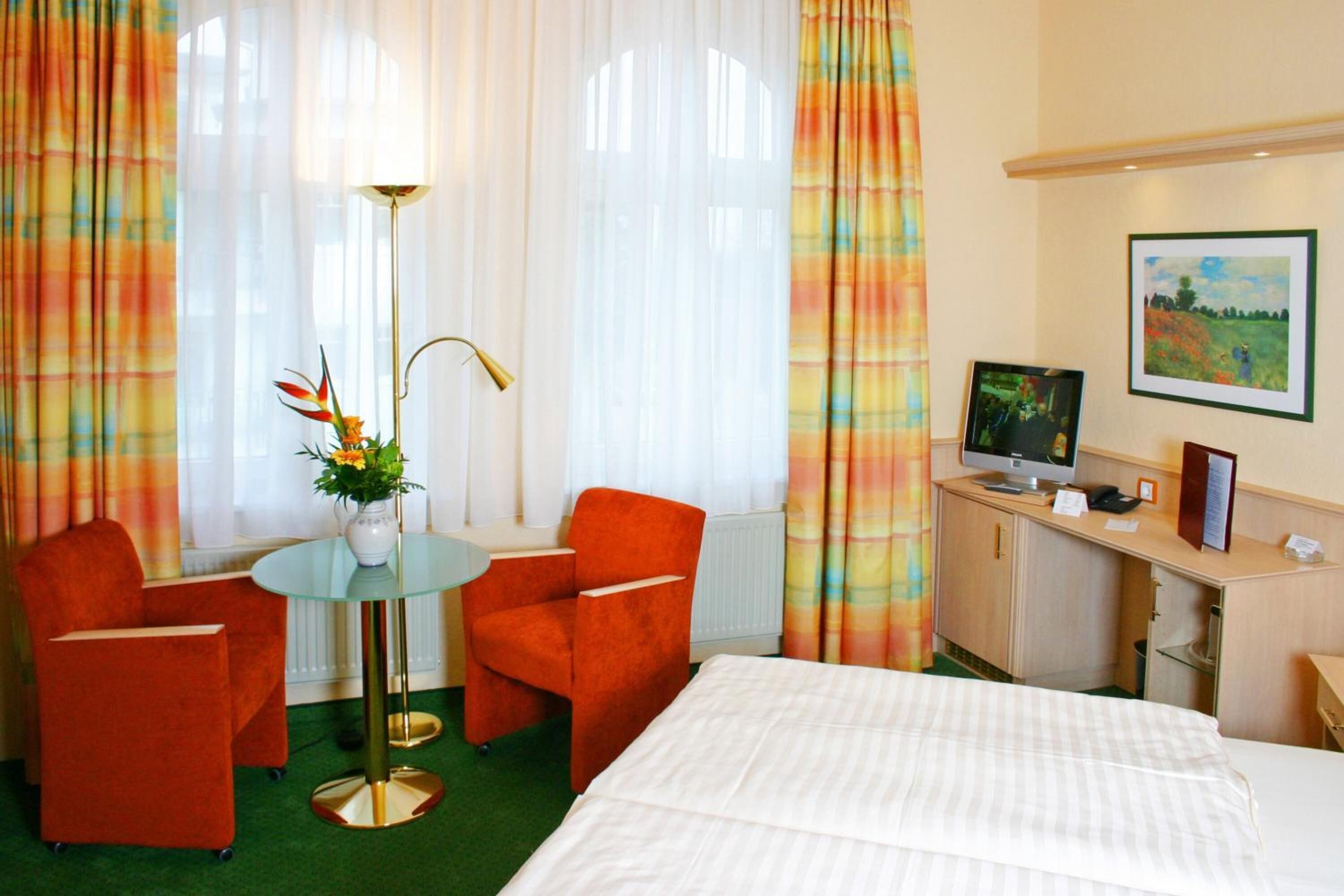 A hotel room with a bed, two red armchairs, a small round table with a vase of flowers, a desk with a computer, and a painting on the wall.