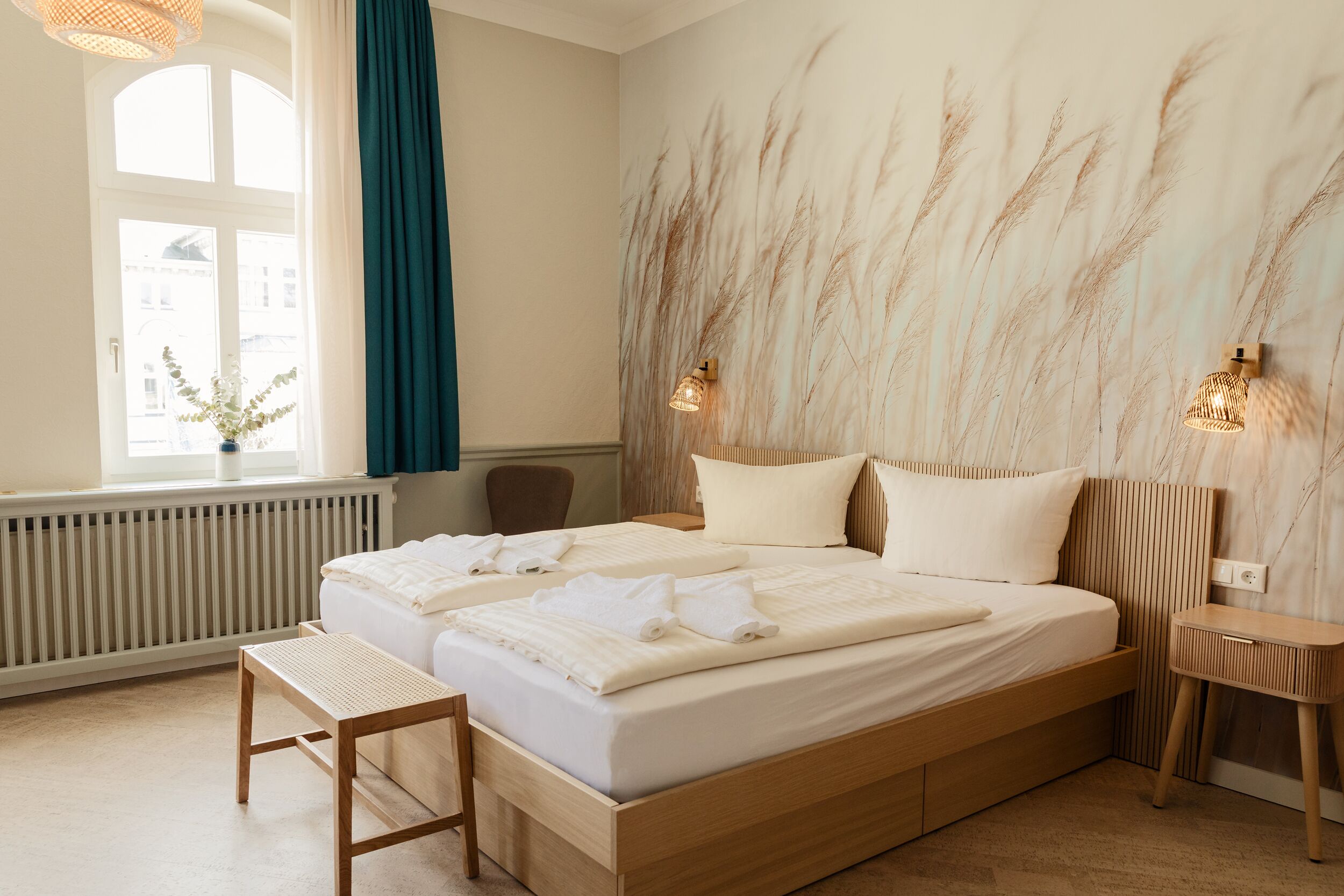 Ein Hotelzimmer mit einem großen Bett, einem Nachttisch aus Holz, einem kleinen Nachttisch, einem Fenster mit Vorhängen und einer Wand mit strukturierter Gestaltung.
