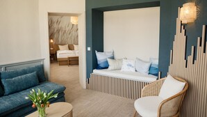 A modern hotel room with a blue sofa, a white chair, a wooden bench with cushions, and a decorative wall panel. - Dünenschloß (Zinnowitz)