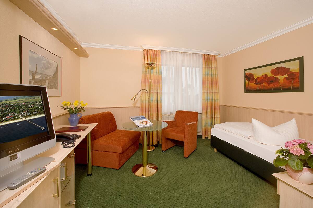 A hotel room with a bed, two armchairs, a small table, a desk with a TV, and a remote control.