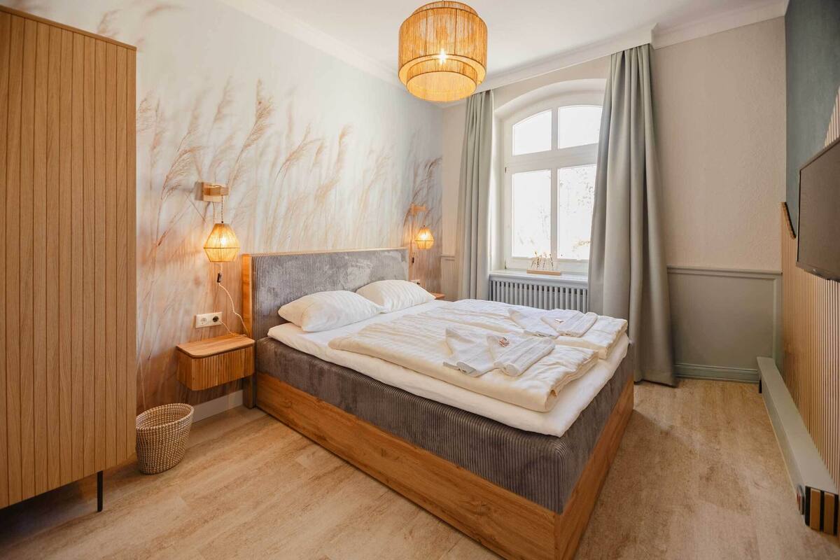 A bedroom with a wooden bed, a nightstand, a wall-mounted lamp, and a window with curtains.