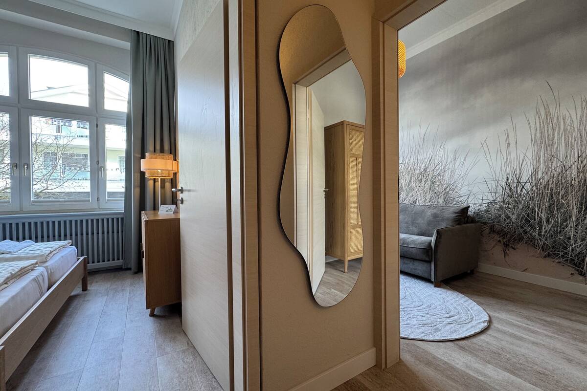 A modern hotel room with a bed, a desk with a lamp, a large mirror, and a view of the outdoors through a window.