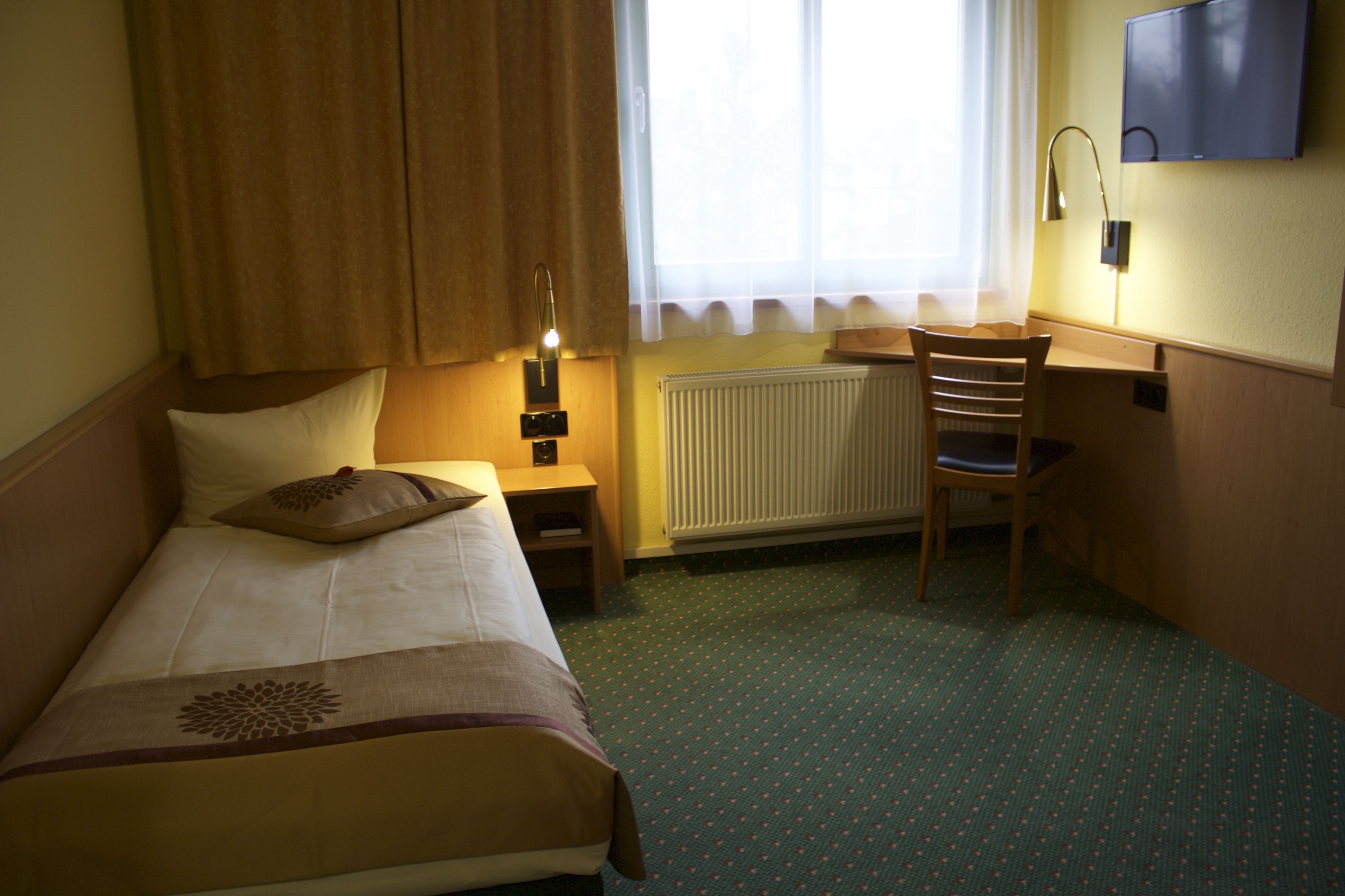 Ein Hotelzimmer mit einem Bett, einem Schreibtisch, einem Stuhl, einem Heizkörper und einem an der Wand befestigten Fernseher.