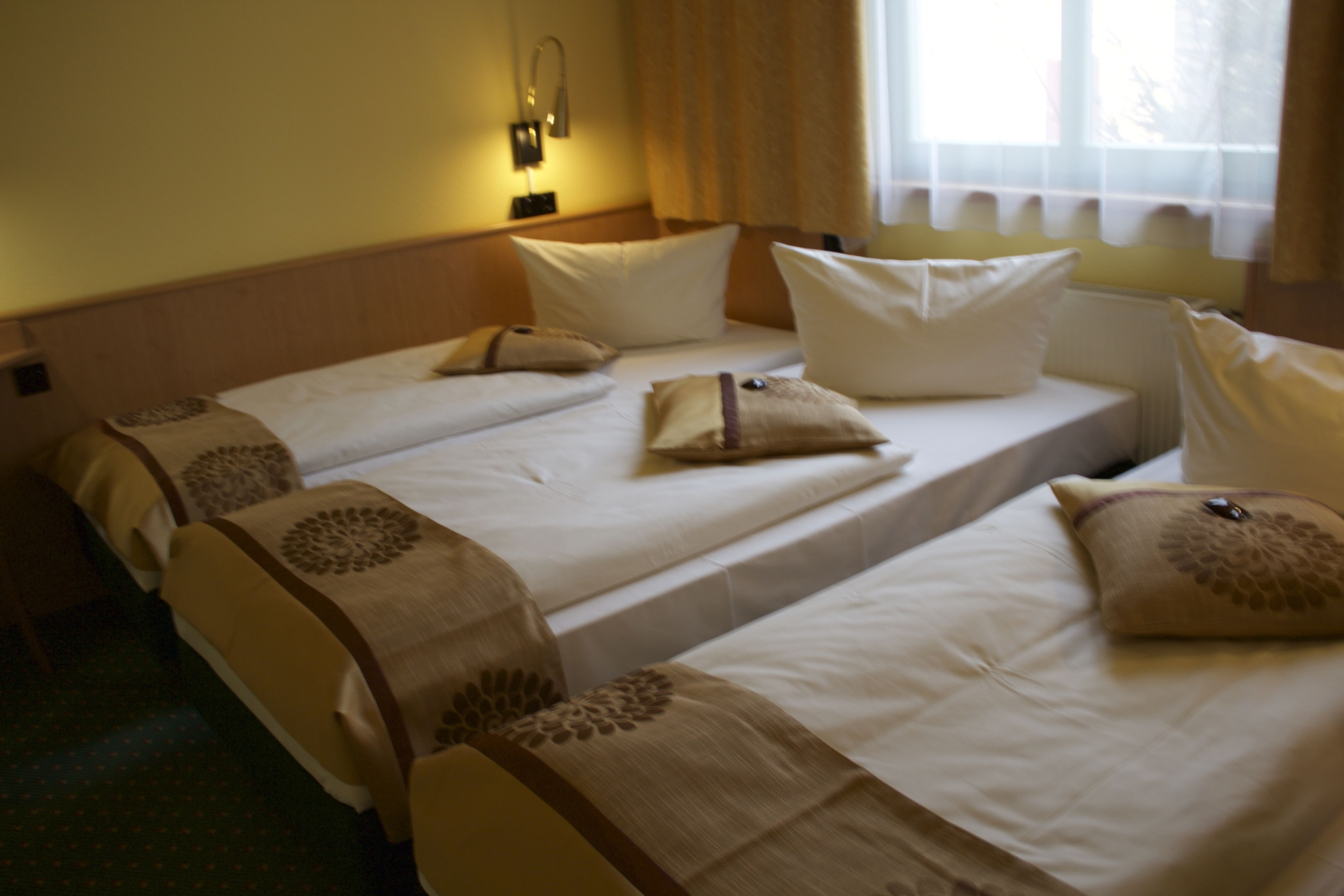 A hotel room with two single beds, each with a pillow and a blanket. There is a nightstand with a lamp and a window with curtains.