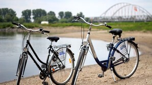 Due biciclette parcheggiate lungo un fiume, con un ponte sullo sfondo.