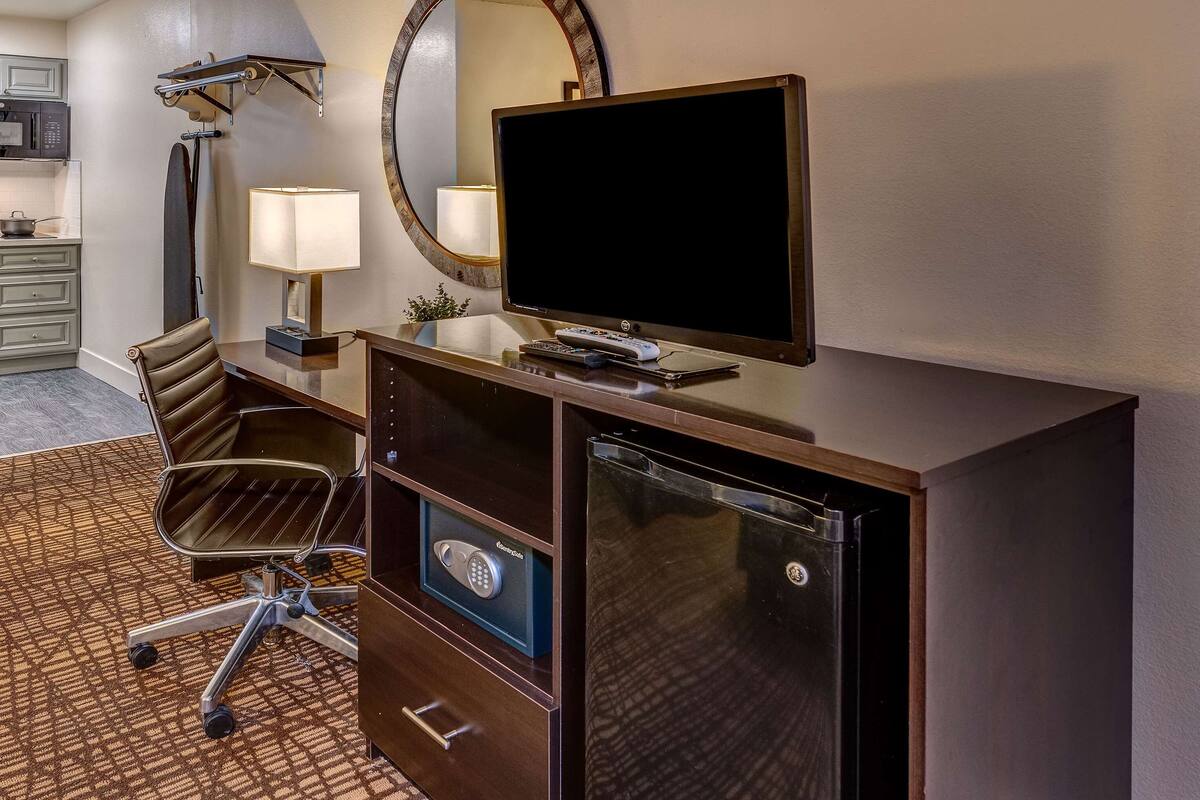 A hotel room with a desk, a flat-screen TV, a lamp, a chair, a microwave, and a refrigerator.
