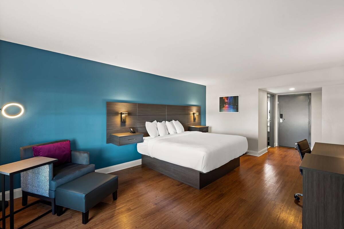 A modern hotel room with a large bed, a desk, a chair, and a small seating area.