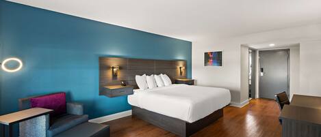 A modern hotel room with a large bed, a desk, a chair, and a small seating area.