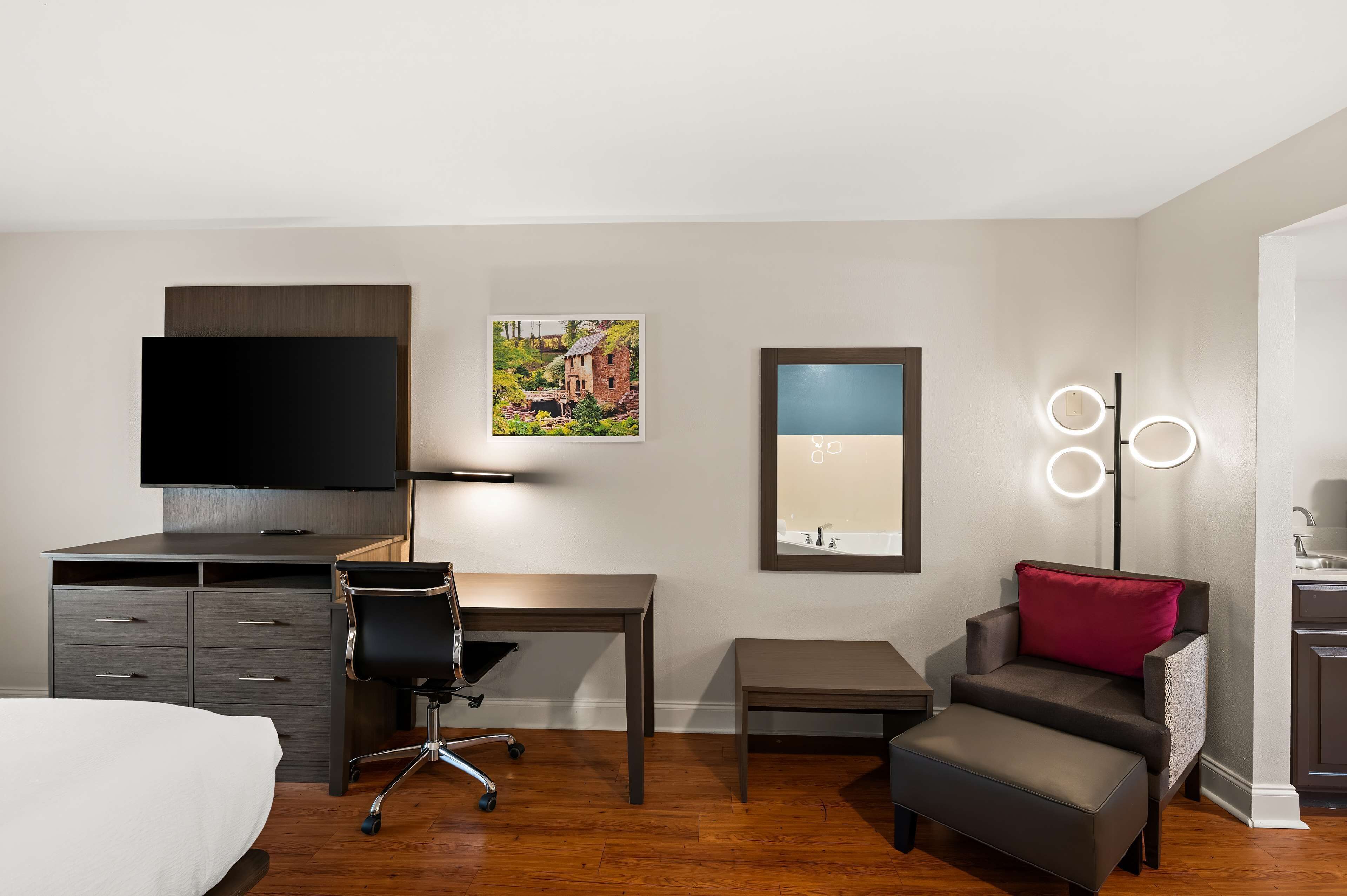 A hotel room with a bed, a desk, a chair, a small table, a sofa with a red pillow, a TV, a picture on the wall, and a mirror with lights.