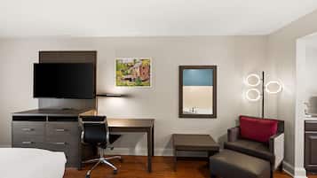 A hotel room with a bed, a desk, a chair, a small table, a sofa with a red pillow, a TV, a picture on the wall, and a mirror with lights.