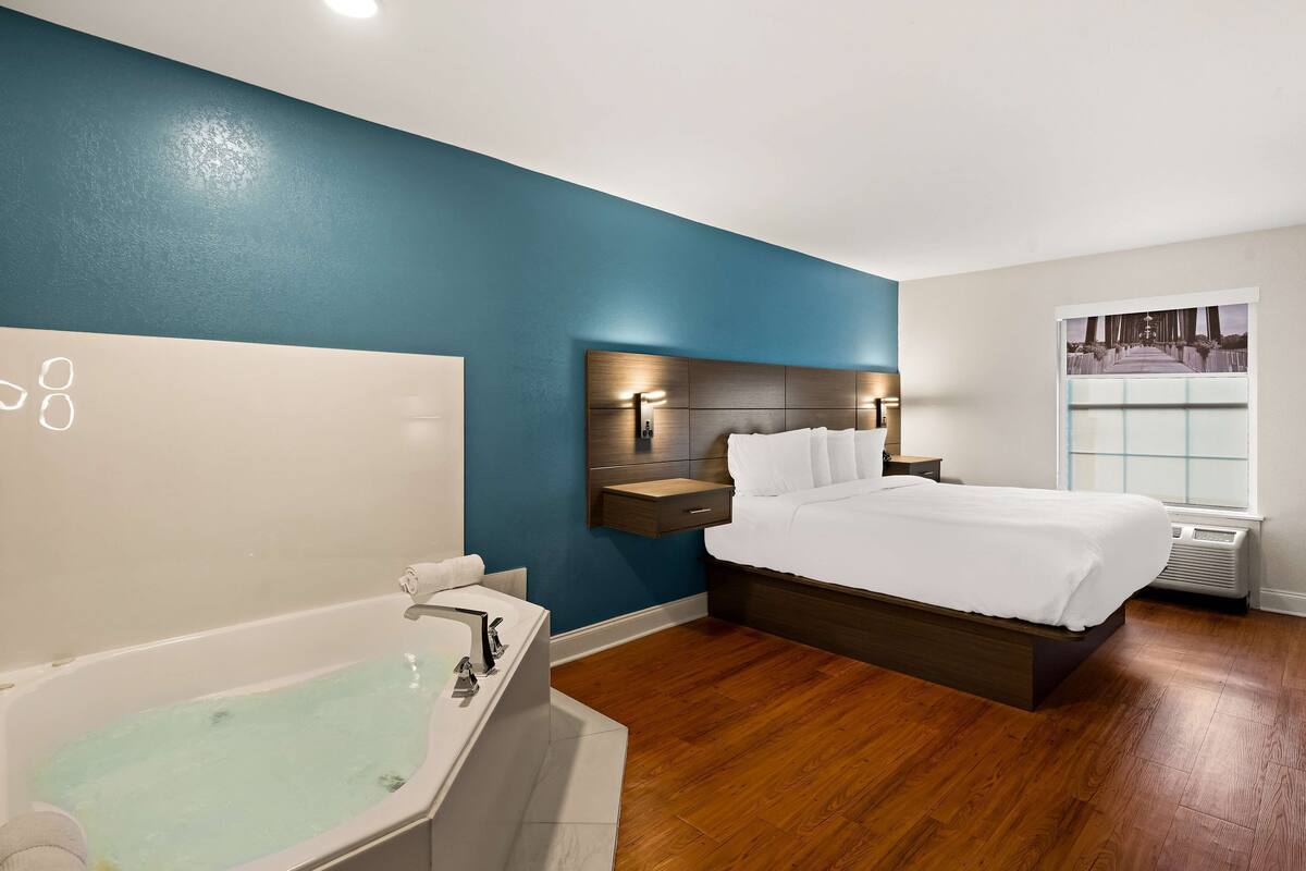 A modern hotel room with a large bed, a built-in nightstand, a wall-mounted light, and a window with a view.