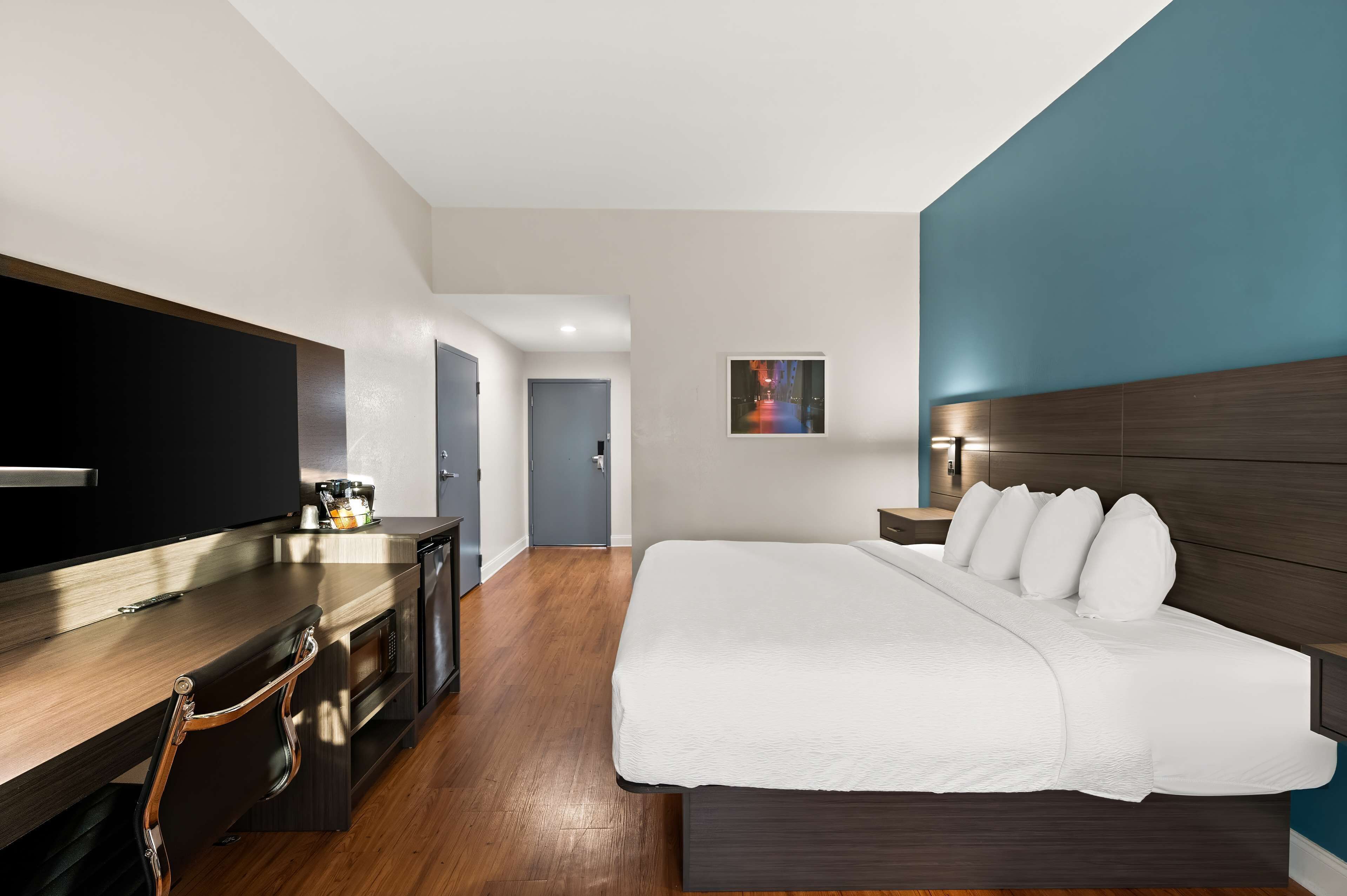 A modern hotel room with a large bed, a flat-screen TV, a kitchenette, and a view of the hallway.