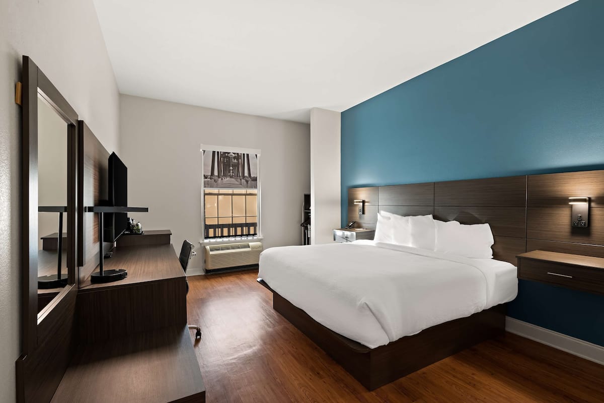 A modern hotel room with a large bed, a desk, a chair, a large mirror, a nightstand, and a wall-mounted lamp.