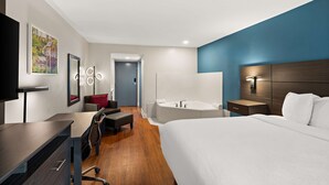 Habitación de hotel moderna con una cama grande, un escritorio, una silla, una lámpara de escritorio, un televisor, un espejo montado en la pared, una puerta y un baño con bañera y ducha.
