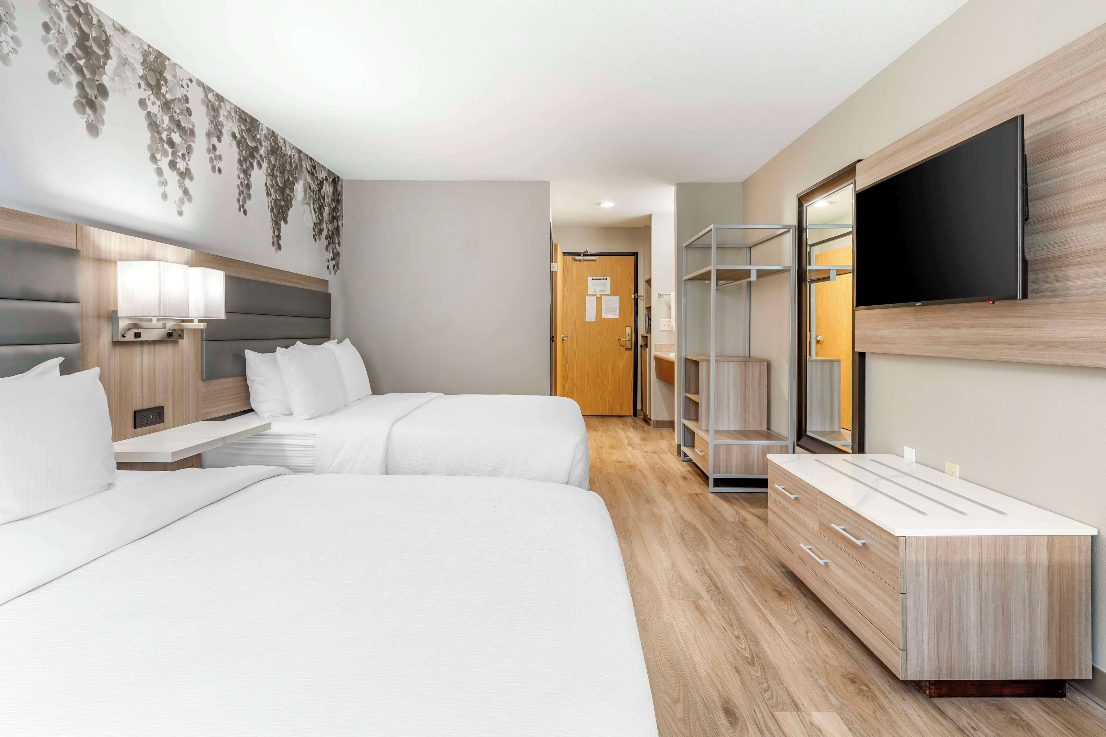 Quarto de hotel moderno com duas camas, uma televisão de tela plana, uma toucador com espelho e uma TV fixada na parede.