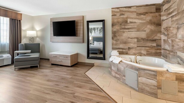 A modern hotel room with a wooden floor, a sofa, a flat-screen TV, a large mirror, a bathtub, and a vanity with a sink. - Best Western Plus Vineyard Inn (Livermore)