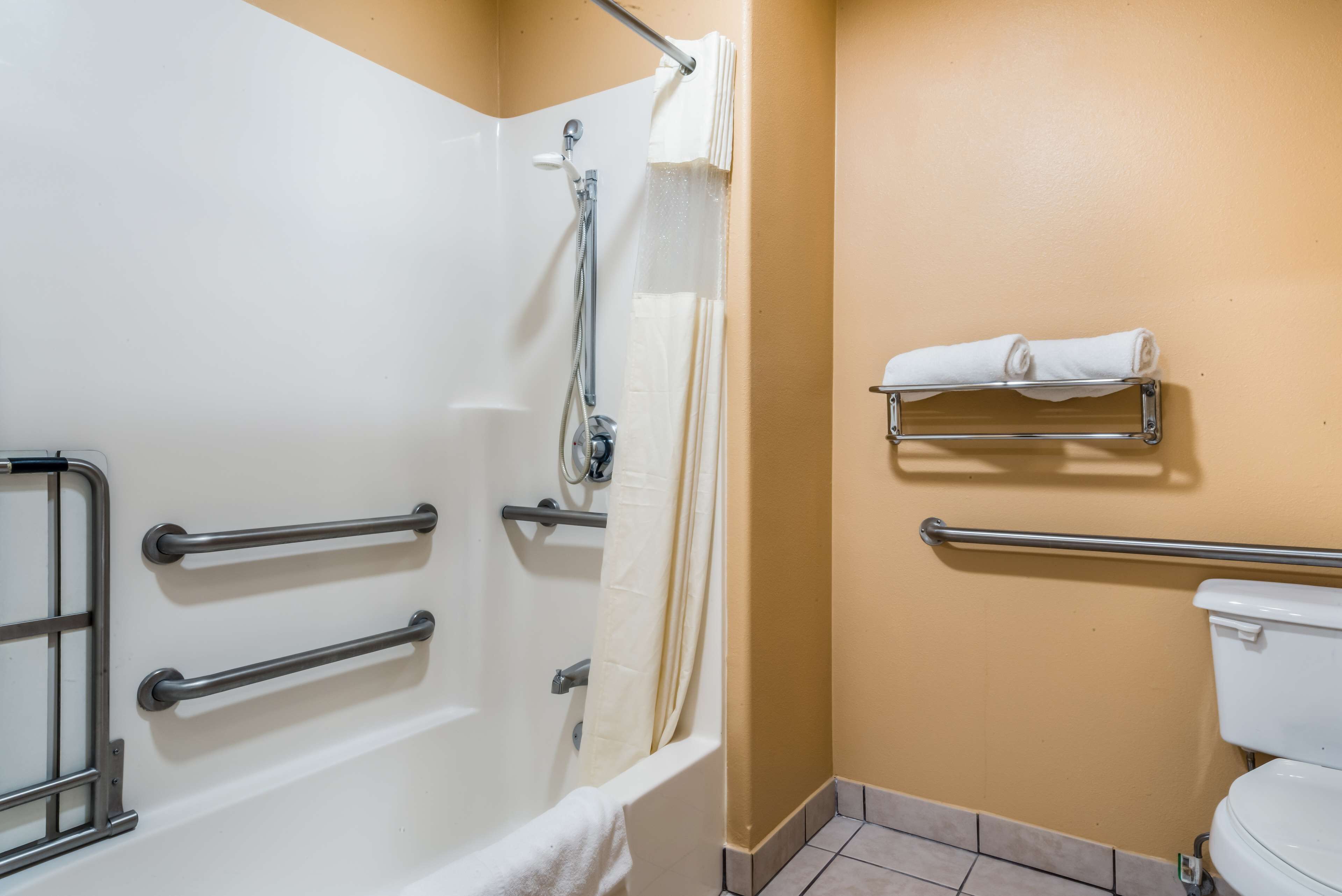 Ein Badezimmer mit Badewanne, Toilette und Dusche. Haltegriffe sind in der Nähe der Badewanne und Toilette angebracht.