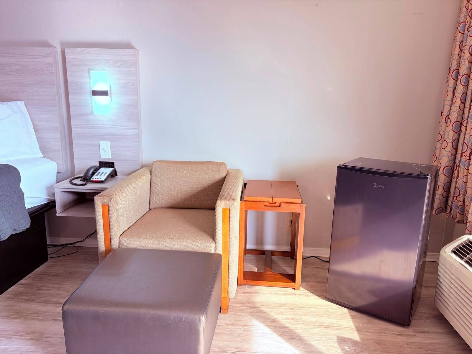 Habitación de hotel con cama, una mesita con teléfono, sofá, mesita de noche de madera, refrigerador y unidad de aire acondicionado.