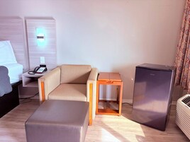 Quarto de hotel com uma cama, uma mesinha com telefone, um sofá, uma mesa de cabeceira de madeira, um frigorífico e um aparelho de ar condicionado.