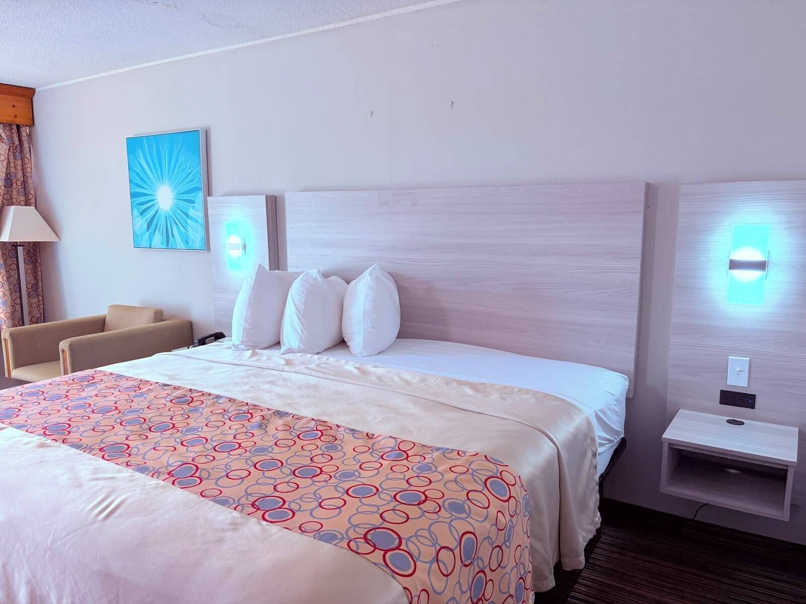 A hotel room with a bed, a bedside table, a lamp, a wall-mounted light, and a painting.
